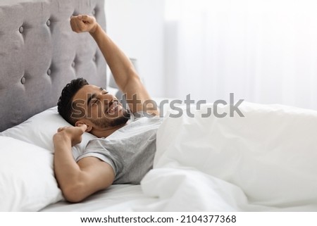 Lazy Morning. Happy Pleased Middle Eastern Man Stretching In Bed After Nice Sleep, Cheerful Young Arab Guy Waking Up With Good Mood, Relaxing In Light Bedroom, Enjoying Weekend Pastime, Copy Space