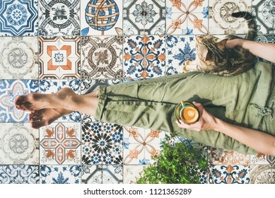 Lazy morning on terrace. Flat-lay of womans legs in cosy linen pants, plant, cat and cup of coffee in hand over colorful moroccan tile floor, top view
