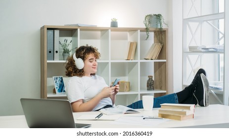 Lazy Student. Homework Break. Chilling Time. Internet Browsing. Happy Sneaky Overweight Curly Hair Woman In Headphones Phone Texting At Modern Light Interior Workplace.