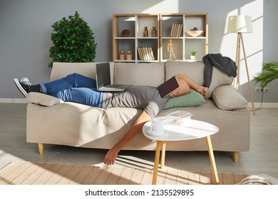 Lazy University Student Falls Asleep While Studying At Home. Tired Young Man Lying On Sofa In Living Room Interior, With Book On Face, Laptop Computer On Tummy And Messy Food Leftovers On Coffee Table