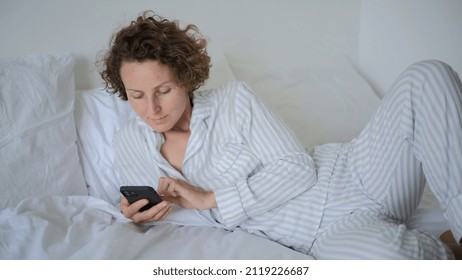 Lazy Weekend Morning, Woman Laying In Bed Scrolling Her Social Media And News Online On The Smartphone