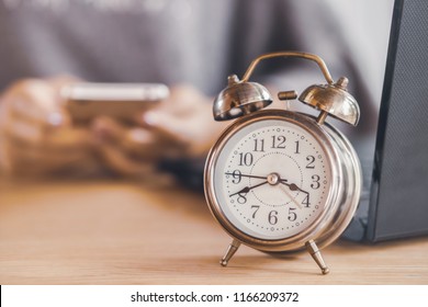 Lazy Woman Worker Wasting Time At Work Using Smart Phone With Alarm Clock Showing The Time On Office Desk