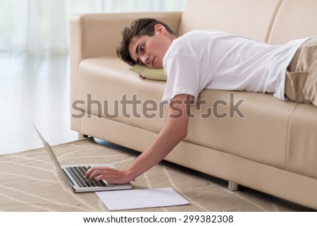 Lazy young man lying on the sofa and watching something on laptop