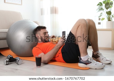 Lazy young man with smartphone eating junk food on yoga mat at home