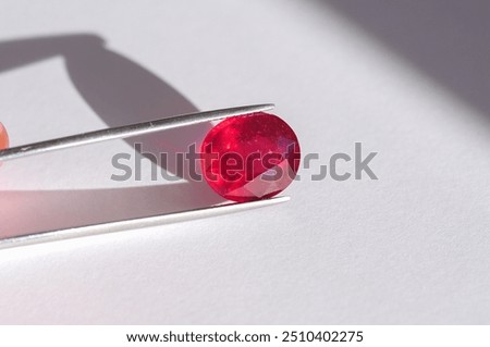 Lead glass filled natural ruby gemstone in tweezers. Heated, treated, affordable red gemstone. Mass market ruby gemstone mined in Madagascar, faceted in Thailand. Bright red colored gemstone setting.