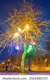 A leafless tree glows with vibrant lights against a dark sky, with a swing set, a person, and a brick building with lit windows in the background.