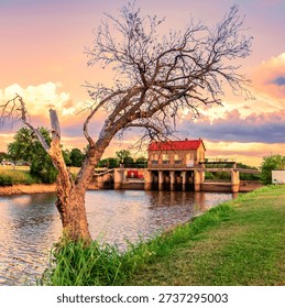 A leafless tree with a twisted, sculptural trunk stands in the foreground of a peaceful riverside scene. Behind it, a small water‑control structure with a red‑tiled roof spans the waterway, its arched openings allowing a steady flow beneath. The grass is lush and green, contrasting beautifully with the dramatic sky above—clouds glowing with warm tones from a rising or setting sun. The blend of natural textures, architectural detail, and vivid sky creates a tranquil, picturesque atmosphere.