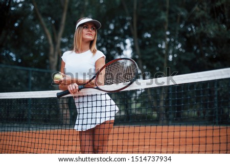 Leans on the net. Female tennis player in sportive clothes on the cort at summer daytime.