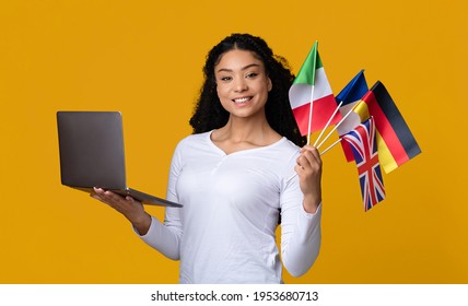 Learn Foreign Languages Online. Portrait Of Happy Black Woman Holding Laptop And International Flags, Young African Female Student Study With Computer, Enjoying Distance Learning, Yellow Background