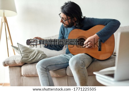 Learn and play. Young caucasian man sitting on sofa at home and adjusting acoustic guitar. Music school online. Distance education. Focus on man. Stay home, self isolation. E-learning