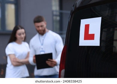Learner driver and instructor with clipboard near car outdoors, selective focus on L-plate. Driving school