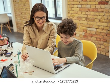 Learning To Code. Smiling Young Female Teacher In Glasses Showing Scientific Robotics Video To Little Boy, Using Laptop In Classroom