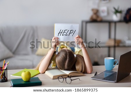 Learning Difficulties, Education Concept. Sad tired frustrated schoolgirl sitting at the table with book and laptop, holding paper with word Help. Overworked child asking for rest. Homeschooling