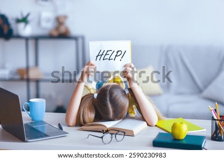 Learning Difficulties, Education Concept. Sad tired frustrated schoolgirl sitting at the table with book and laptop, holding paper with word Help. Overworked child asking for rest. Homeschooling