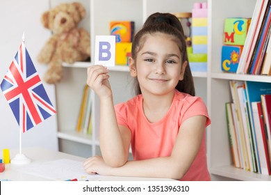 Learning English. Happy Cute Child Girl Sitting At A Table And Draws With A White Bookcase, Colored Books And Toys And British Flag Behind. Back To School And Happy Time! 