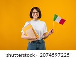 Learning Italian, student exchange and travel. Happy lady holding notebooks and small flag of Italy, ready to study in college, standing over orange background, studio shot