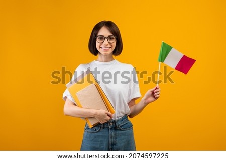 Learning Italian, student exchange and travel. Happy lady holding notebooks and small flag of Italy, ready to study in college, standing over orange background, studio shot
