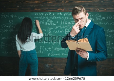 Learning math enables me to think clearly. Teacher man in glasses read problem statement from textbook to girl student. Rear view woman write equation on chalkboard. Doing sums. School day.
