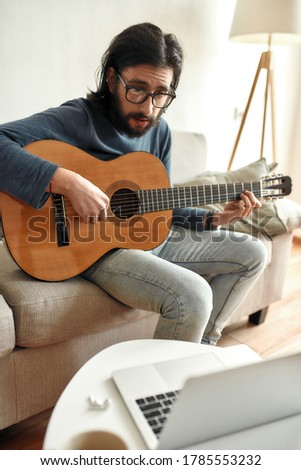 Learning a new instrument. Young focused man wearing glasses sitting on sofa at home and learning how to play guitar, watching online course on laptop. Distance education. Focus on a man. Stay home