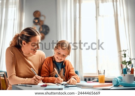 Learning new together. Two generations of optimistic females sitting at corner of desktop and writing in copybooks. Easygoing lady looking at kid doing exercises and pretending practising same task.