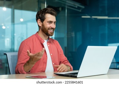 Learning online. A young male teacher in a red shirt sits in the office at the table, works on a laptop, teaches through a video call, conference, webinar. Explains, smiles. - Powered by Shutterstock