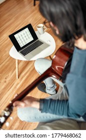 Learning In Self Isolation. Rear View Of Young Man Sitting On Sofa At Home And Learning How To Play Guitar, Watching Online Course On Laptop. Distance Education. Focus On Screen. Stay Home. E-learning