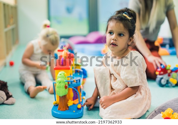 Learning through play at the nursery school. Toddler\
little girl and the teacher playing with colorful plastic\
playhouses, building blocks, cars and boats. Imagination,\
creativity, fine motor\
and