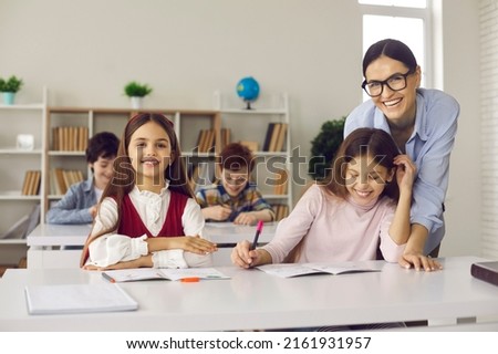 Learning via encouragement. Positive smiling young caucasian teacher and elementary school girl pupil sitting at desk and laughing in classroom. Portrait shot with boy classmates on blurred background