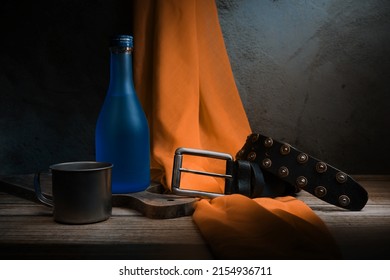 Leather Belt, Blue Bottle And Drinking Cup On The Table. Fine Art Photography. Light Painting Photography. Still Life Photography.                               