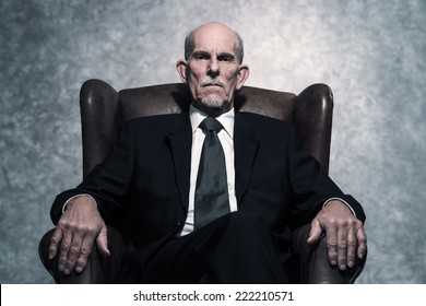 In Lederstuhl sitzend Senior Business Mann mit grauem Bart mit dunklem Anzug und Krawatte. Gegen graue Wand.