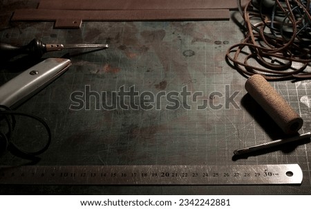 Leatherwork requires tools laid out on a worn cutting board. Cutting blade, edge beveler, cork, ruler, hole puncher. Twisted leather strips. Dark background. Copy space. Minimal lamp lighting