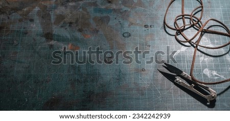 Leatherwork requires tools laid out on a worn cutting board. Ruler, scissors. Twisted leather strips. Dark background. Copy space. Minimum lamp illumination