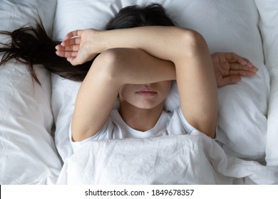 Leave me alone! Top view of capricious girl teenager or young female lying in cozy bed resisting unwilling to wake up early in the morning hiding face under crossed hands covering eyes from sunlight