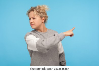 Leave me! Resentful woman with curly hair in sweatshirt pointing sideways, showing get out gesture, ordering to go away, feeling annoyed and vexed. indoor studio shot isolated on blue background