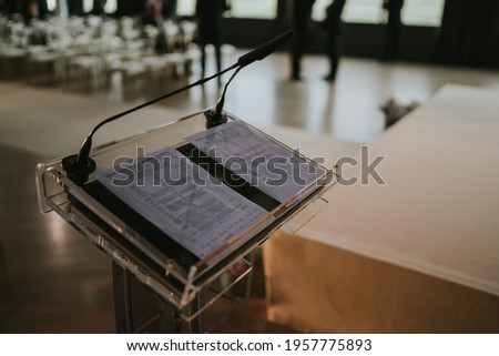 LECTERN WITH TWO MICROS AND AN OPEN BOOK WITH TEXT IN HEBREW IN EMPTY ROOM FULL OF CHAIRS