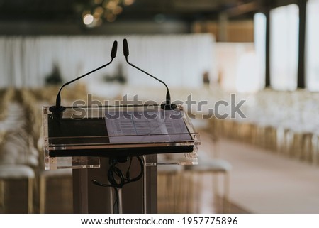 LECTERN WITH TWO MICROS AND AN OPEN BOOK WITH TEXT IN HEBREW IN EMPTY ROOM FULL OF CHAIRS
