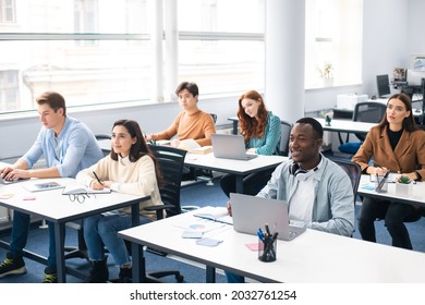 Lecture Concept. Group of interested multicultural mixed race group of students sitting at tables with pc in modern classroom, listening to teacher, taking notes writing in notebooks, back to study