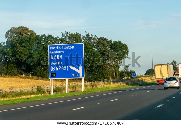 Leeds, UK - 23 August 2019: Blue British Motorway\
sign advising drivers on the A1 to take Junction 51 to\
Northallerton, Leyburn and Bedale. Traffic concept, highway,\
freeway, rising fuel\
price.