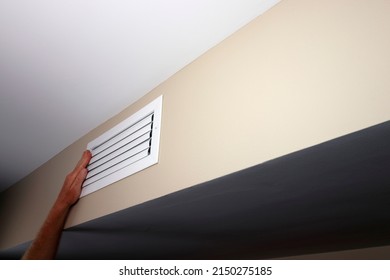 A left caucasian adult hand held up to a closed small air vent on a beige wall near a white ceiling. White metal rectangle furnace closed vent grid with a left hand on top of the left side close-up