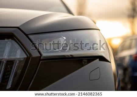 Left front full LED headlamp of modern black electric crossover SUV, dusk light in the background, blurred background, other cars behind. Part of grille, bonnet and bumper with lamp washer visible