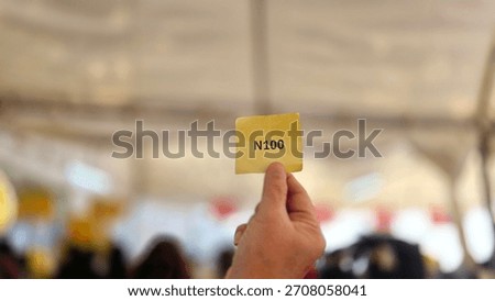 Left hand holding queue ticket number N100 printed in black ink on yellow paper with blur background. Queues for transactions in banks, government agencies, hospitals. Will be served in the 100th orde