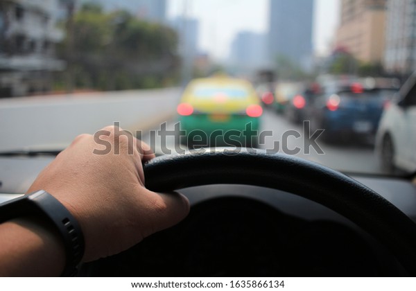 The left hand of a
man driving a car  With the left hand holding the steering wheel
and the wrist, hand, black watch as well  The ground is a blurry
image of a traffic blur.