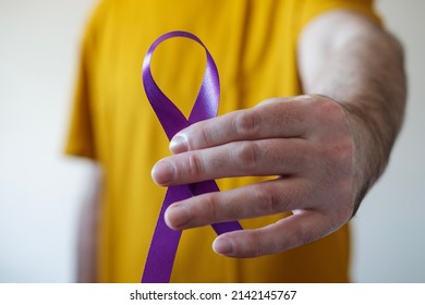 Left hand man holding a purple ribbon. Yellow t-shirt. Epilepsy, pancreatic cancer and Alzheimer's symbol.