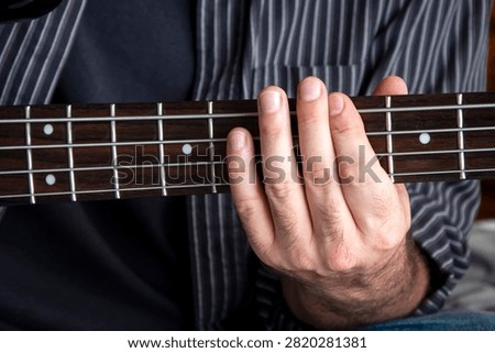 Musician’s left hand positioned on the neck of an electric bass guitar, pressing the strings to form notes and chords. The image emphasizes finger placement, fretboard details, and the expressive technique used in bass performance. Ideal for themes of music, musicianship, practice, learning, recording, live performance, bands, and musical instruments.