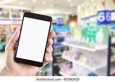 Left Hand Using Smartphone With Blank Screen On Abstract Blur Background Inside Pharmacy Store With Shelves Of Pharmaceutical And Drug.