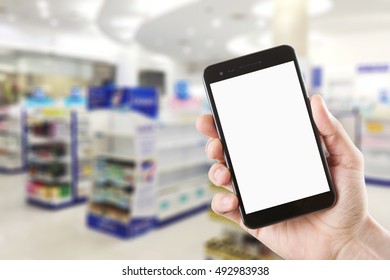 Left Hand Using Smartphone With Blank Screen On Abstract Blur Background Inside Pharmacy Store With Shelves Of Pharmaceutical And Drug.