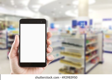 Left Hand Using Smartphone With Blank Screen On Abstract Blur Background Inside Pharmacy Store With Shelves Of Pharmaceutical And Drug.