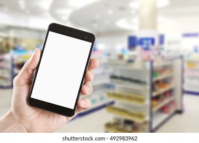 Left Hand Using Smartphone With Blank Screen On Abstract Blur Background Inside Pharmacy Store With Shelves Of Pharmaceutical And Drug.