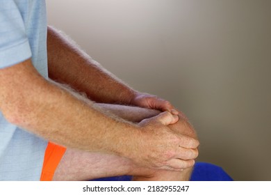 Left knee being self massaged by a mature caucasian man wearing orange sports clothes blue shirt close-up. Massaging to relieve pain felt in his left knee an adult white male massages with both hands