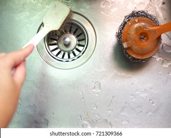 Left Lady Hand Using Plastic Iron Mesh Brush Scrubbing Cleaning Wet Stainless Steel Basin Tub, With Blurred Orange Scratching Tool And Water Drain Background